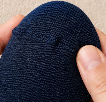 Macro close-up of the flat toe seam on a navy Kilde mercerized cotton sock held between two fingertips on beige linen